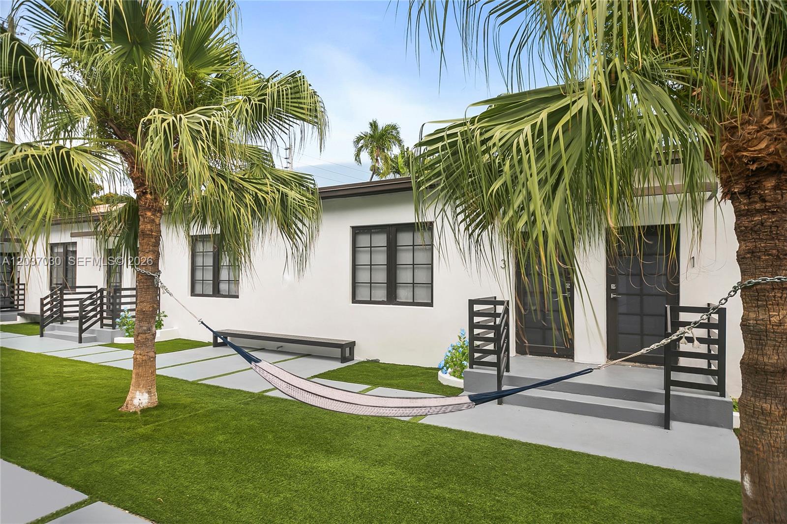 A hammock hangs between two palm trees in front of a white apartment building — MIAMI BEACH, FL