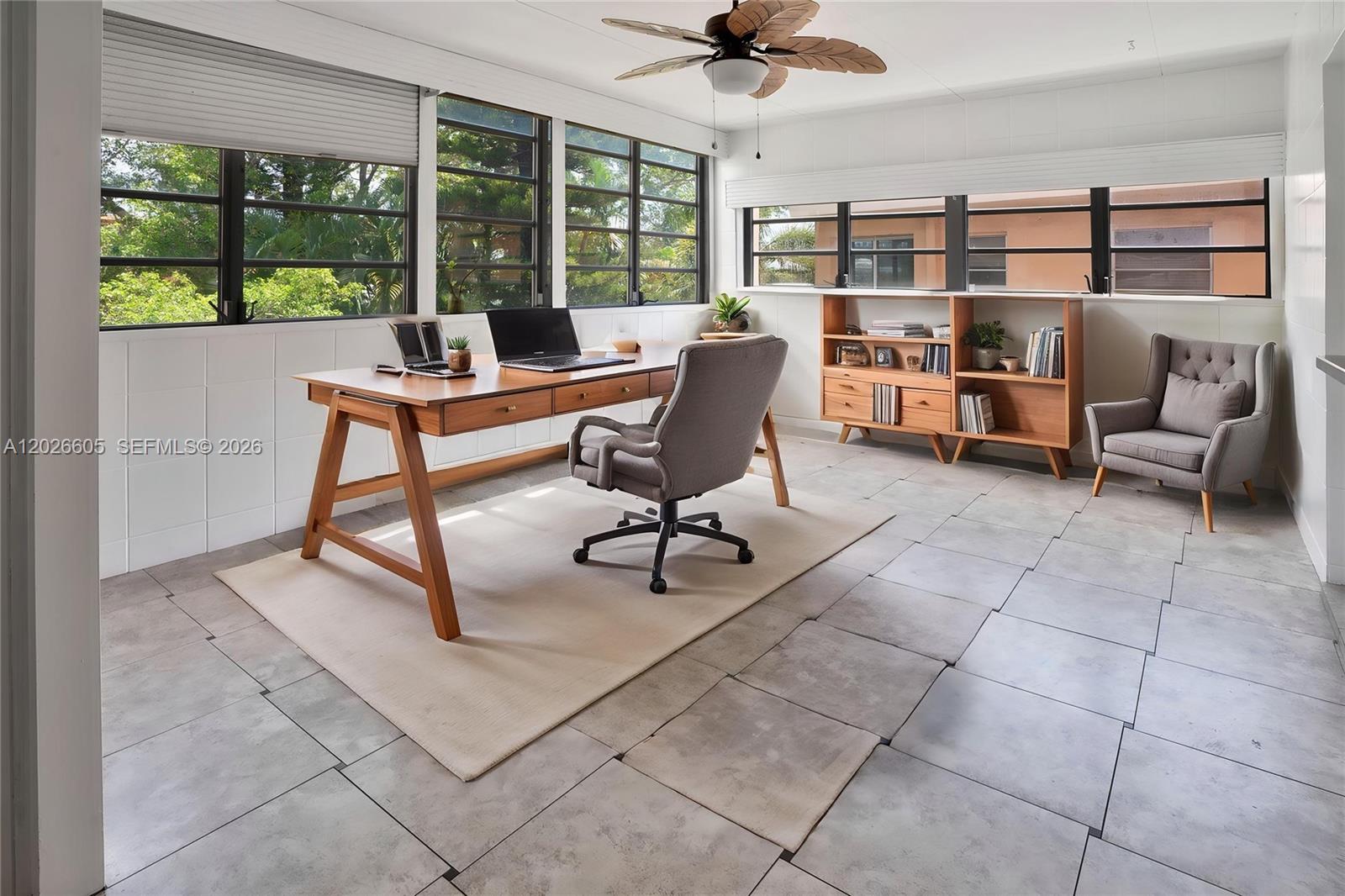 A home office features a wooden desk, gray chair, bookshelf, and large windows with greenery — 180 NE 12th Ave # 18C, Hallandale Beach FL 33009, HALLANDALE BEACH, FL