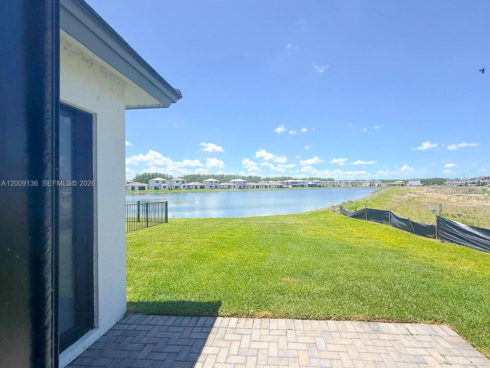 A paved patio overlooks a grassy backyard, a lake, and distant houses under a blue sky — 5639 Argento Drive # 5639, Ave Maria FL 34142, AVE MARIA, FL