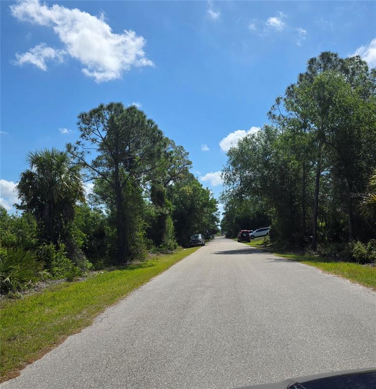 A paved road stretches into the distance, flanked by dense trees and vegetation under blue skies — 23445 CHIMES AVENUE, PUNTA GORDA, FL