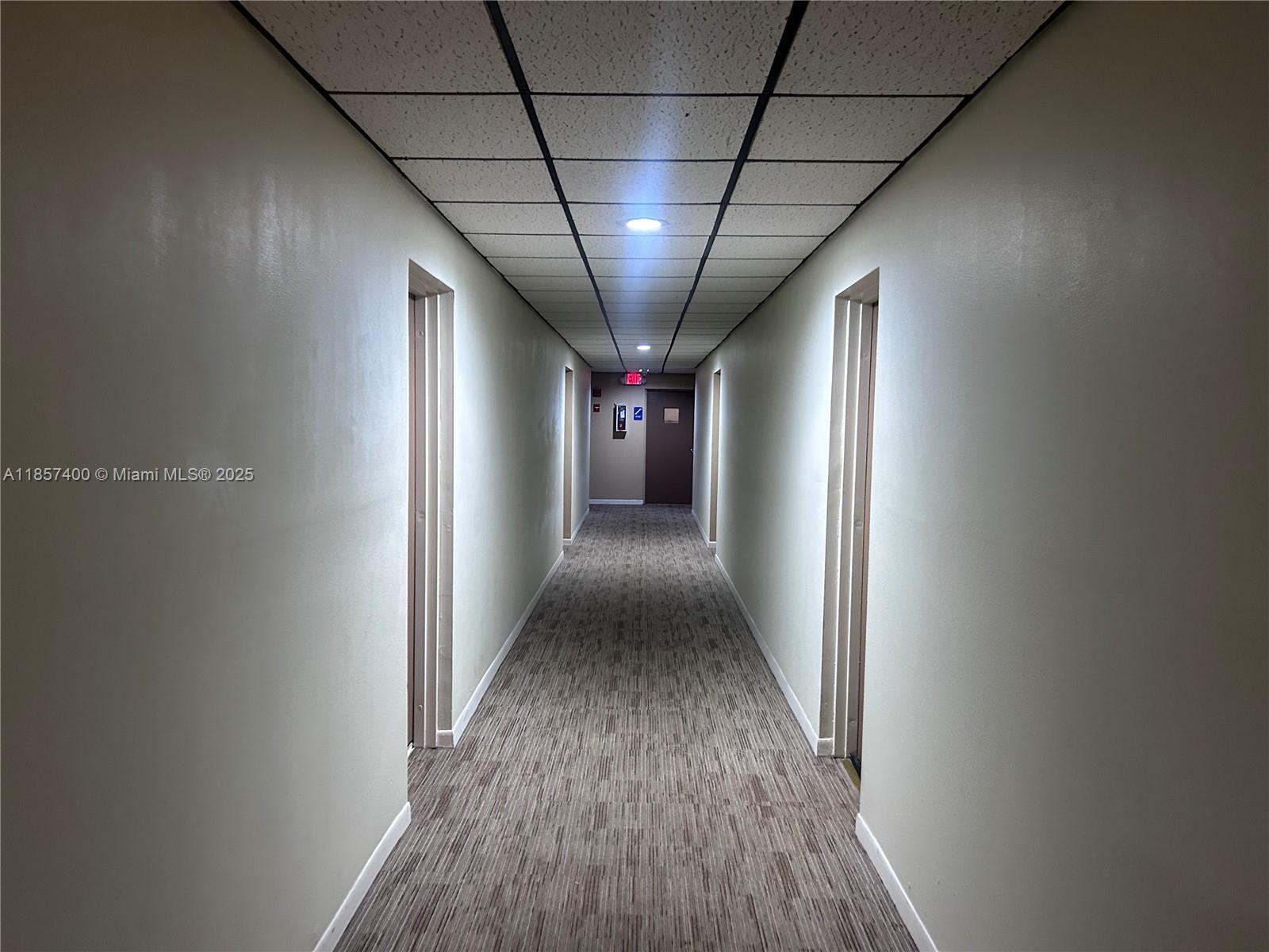 A long, narrow indoor hallway features beige walls, a drop ceiling, and gray carpeted flooring — 14201 SW 66th St # 306, Miami FL 33183, MIAMI, FL