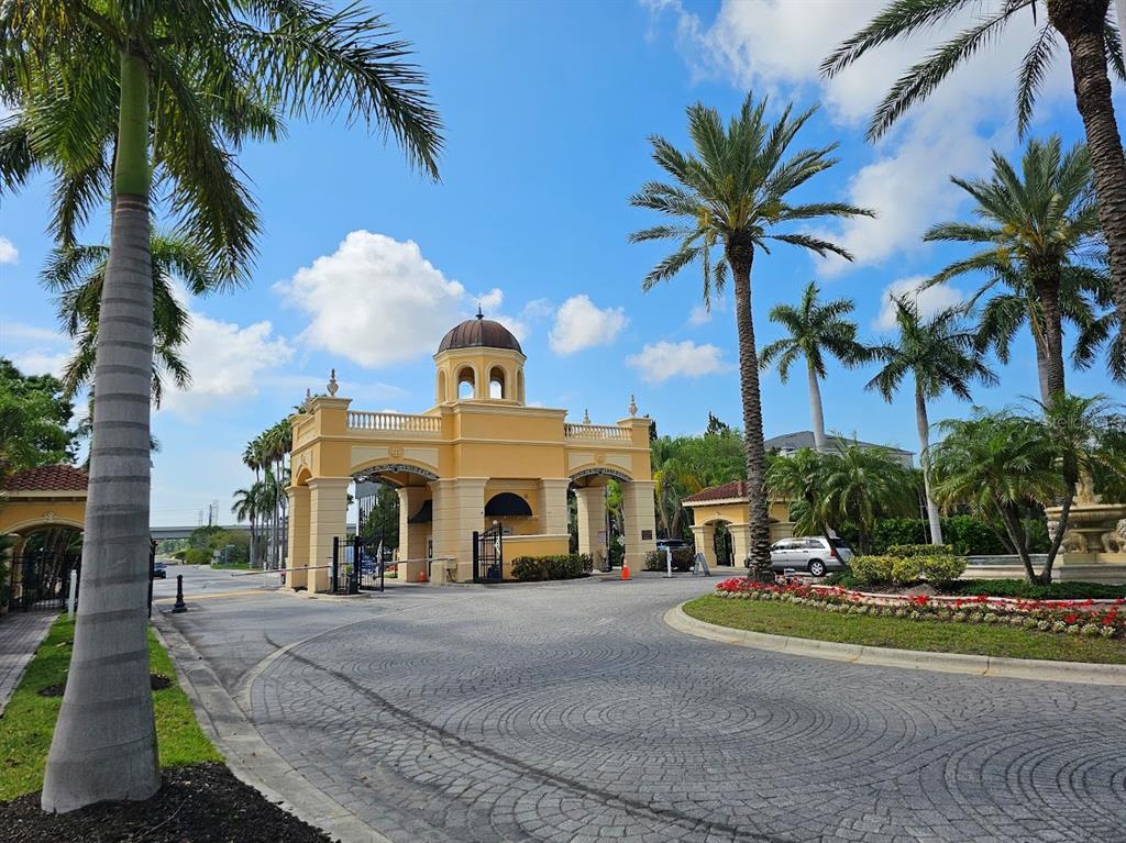 A gated entrance with a yellow stucco guardhouse and palm trees under a blue sky — 2713 VIA MURANO UNIT 236, CLEARWATER, FL