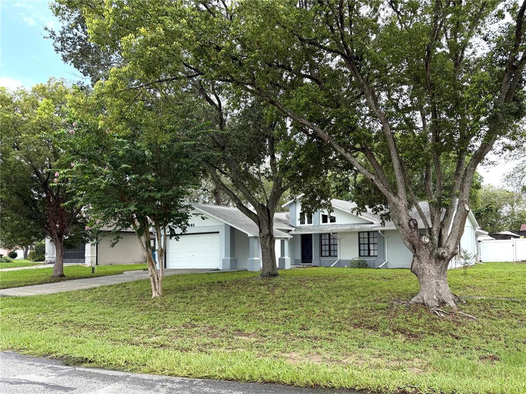 A single-story house with a white garage and gray siding sits behind large mature trees — 7463 PHILATELIC DRIVE, SPRING HILL, FL