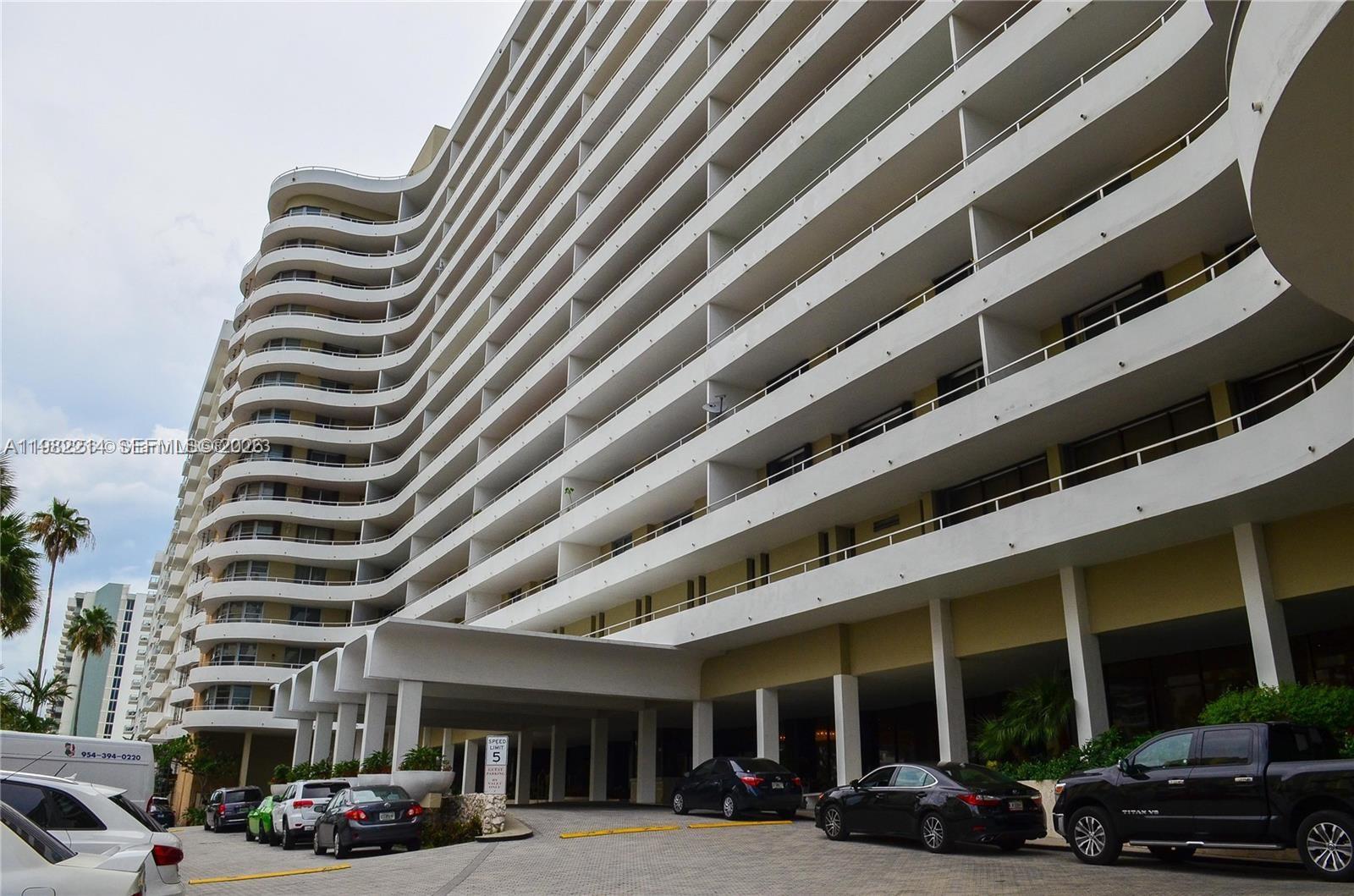 A low-angle exterior view of a large, multi-story white apartment building with curved balconies — 5555 Collins Ave # 4A, Miami Beach FL 33140, MIAMI BEACH, FL