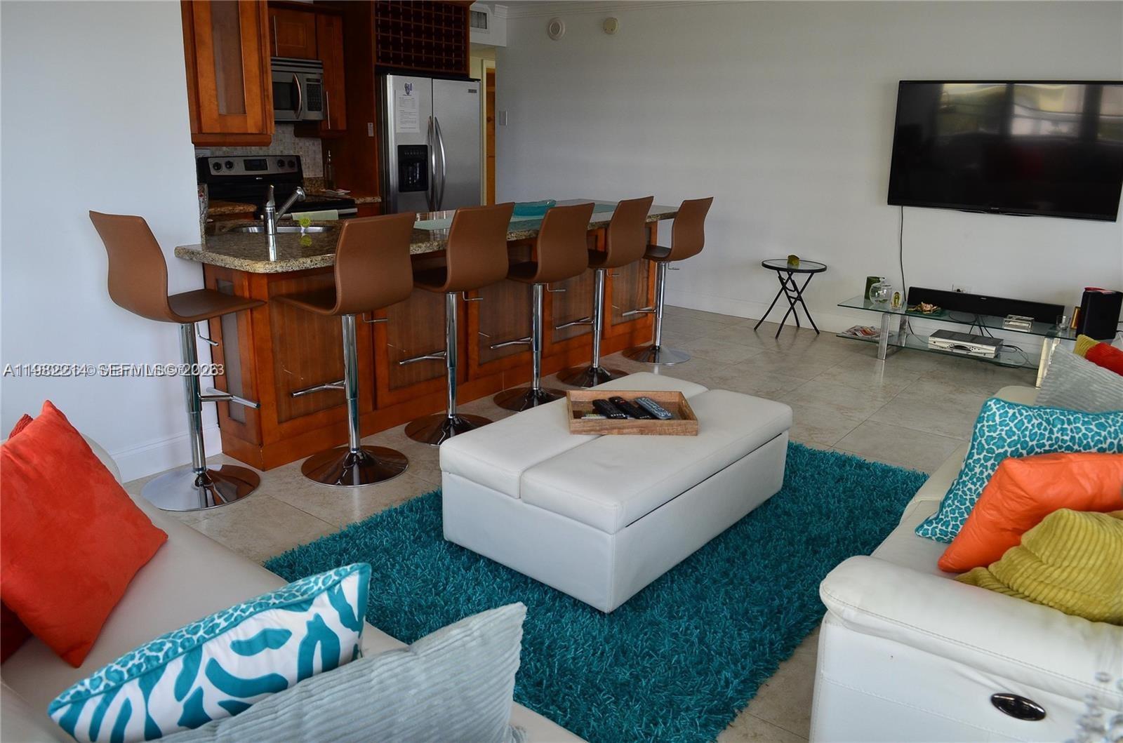 An open-concept living room features a kitchen island with bar stools and a white ottoman — 5555 Collins Ave # 4A, Miami Beach FL 33140, MIAMI BEACH, FL