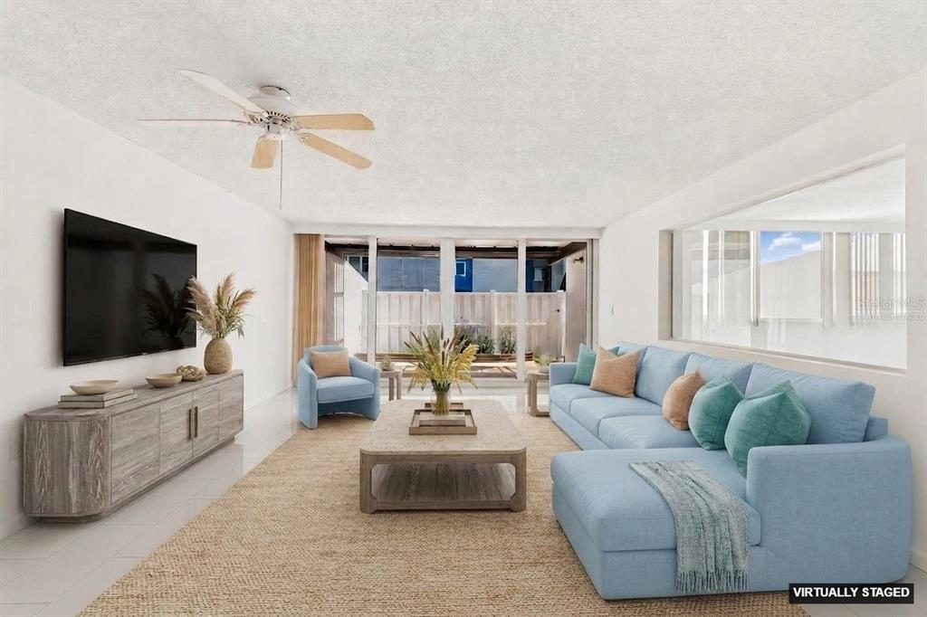 A virtually staged living room features a blue sectional, light wood console, and sliding glass doors — 450 TREASURE ISLAND CAUSEWAY UNIT 106, TREASURE ISLAND, FL