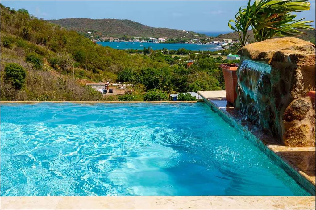 An infinity pool with a stone waterfall feature overlooks a bay and hillside landscape — 5R MOUNT RESACA BARRIO FLAMENCO, CULEBRA, PR