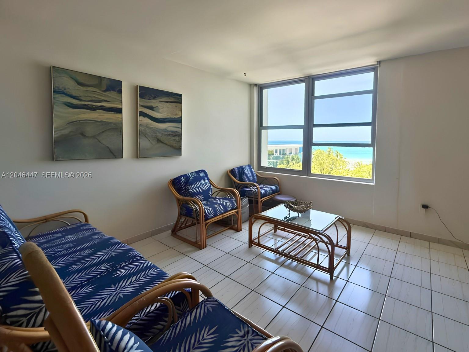 A living room features rattan furniture with blue patterned cushions, tile flooring, and ocean views — 2899 Collins Ave # 905, Miami Beach FL 33140, MIAMI BEACH, FL