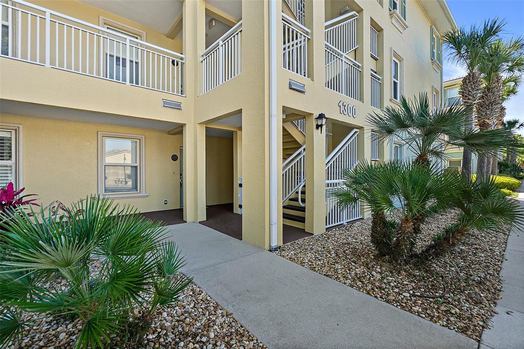 A yellow multi-story apartment building exterior features a concrete walkway, stairs, and palm trees — 1300 CANOPY WALK LANE UNIT 1314, PALM COAST, FL