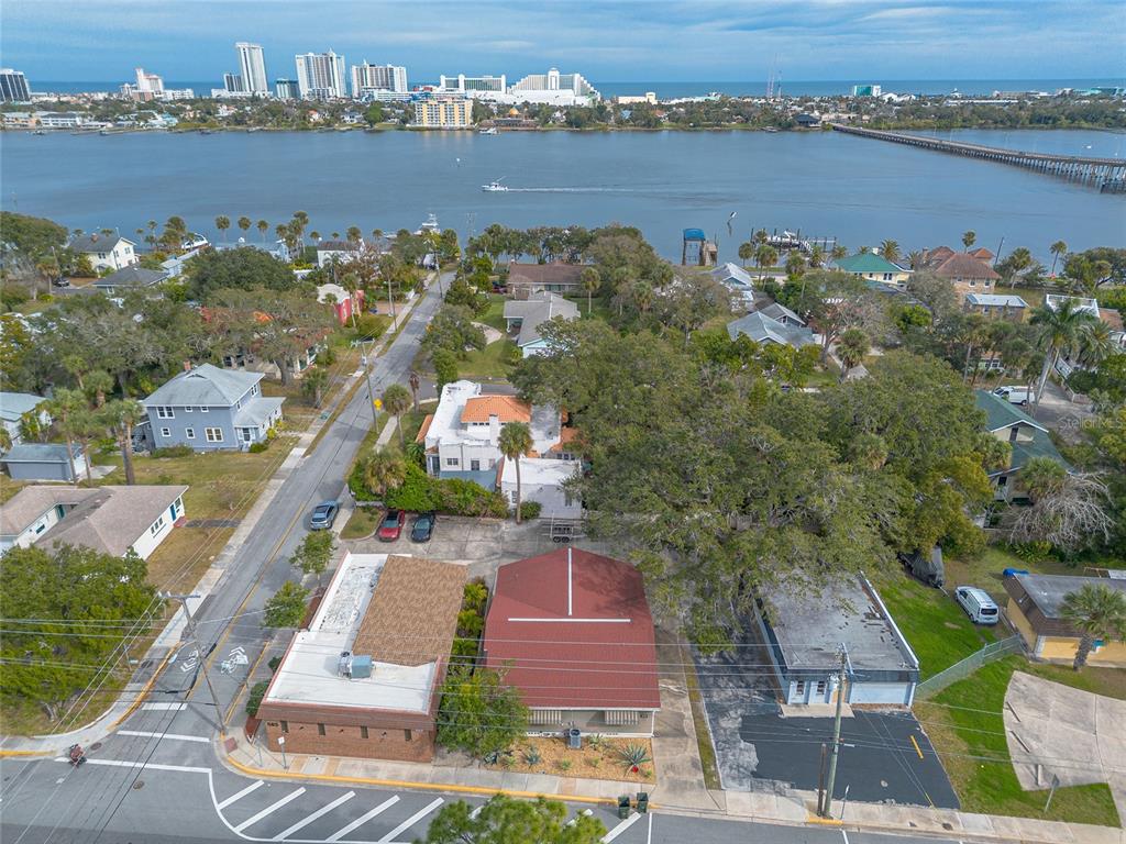 An aerial view shows commercial buildings and houses near a river with a distant skyline — 555 BALLOUGH ROAD, DAYTONA BEACH, FL