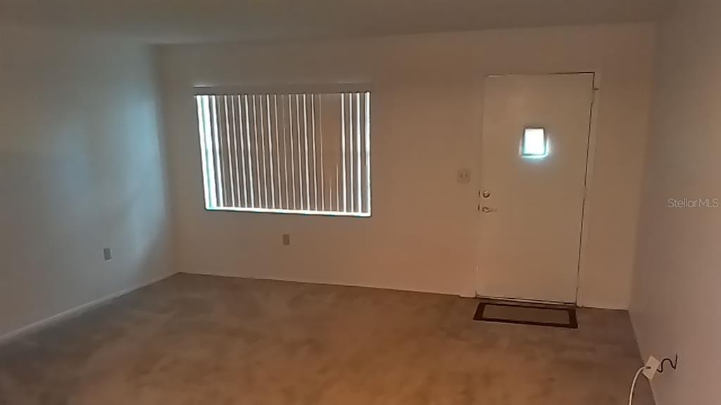 An empty room with beige carpet, white walls, vertical blinds on a window, and a door — 706 GREEN VALLEY ROAD UNIT E4, PALM HARBOR, FL
