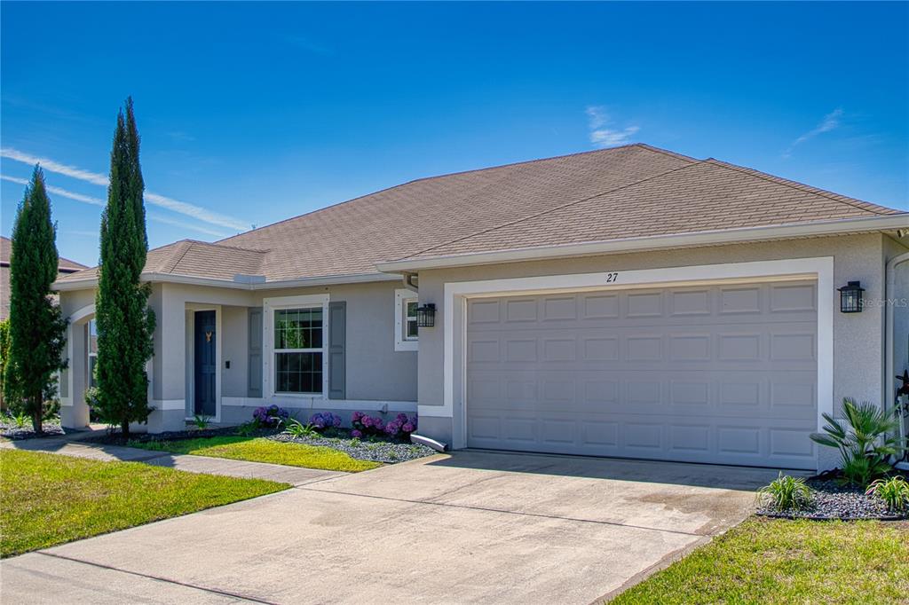 A single-story house with a two-car garage, light-colored stucco exterior, and two tall cypress trees — 27 LEWISDALE LANE, PALM COAST, FL