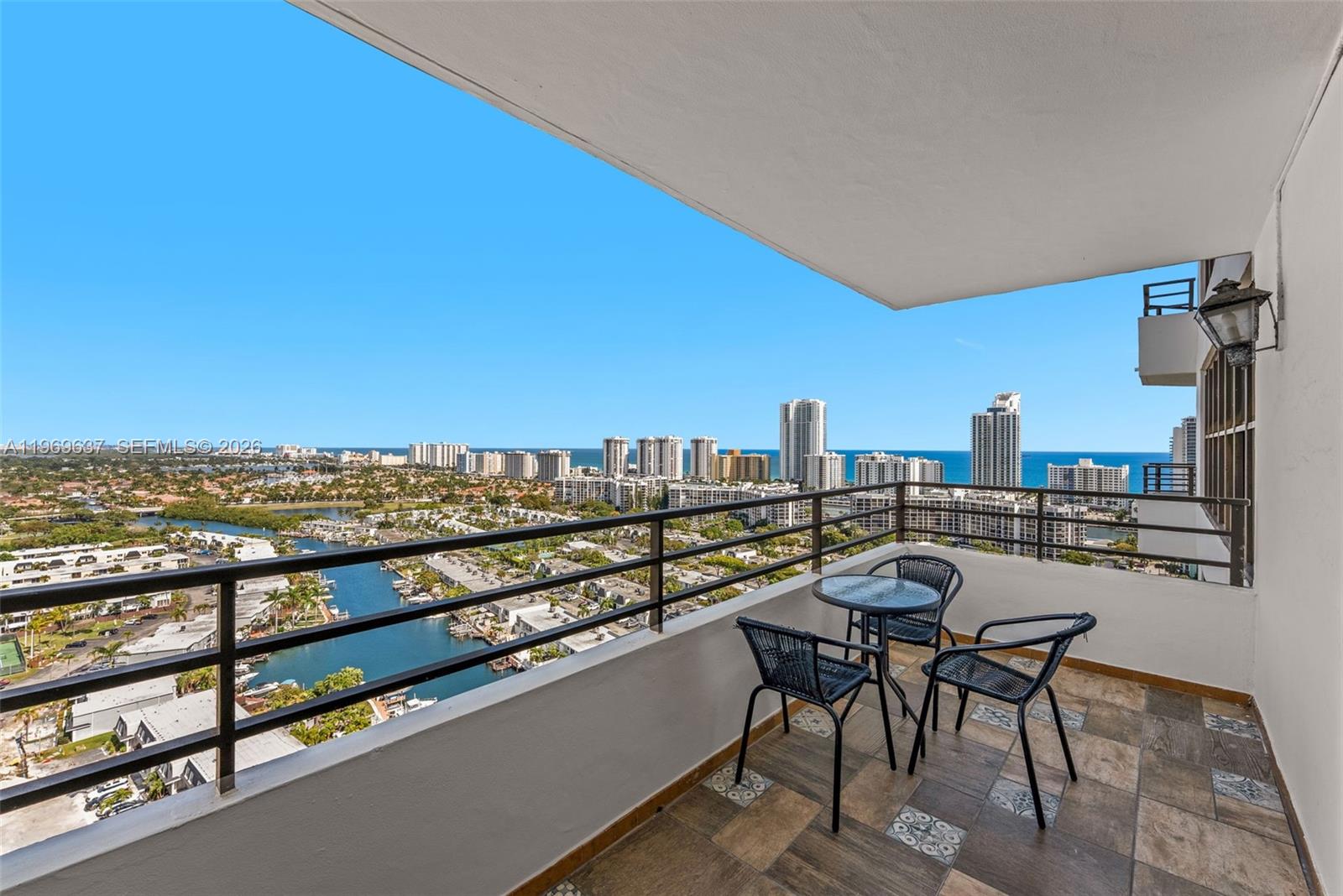 A balcony with tile flooring, a small table, and three chairs overlooks a coastal city — 2500 PARKVIEW DR # 2407, Hallandale Beach FL 33009, HALLANDALE BEACH, FL
