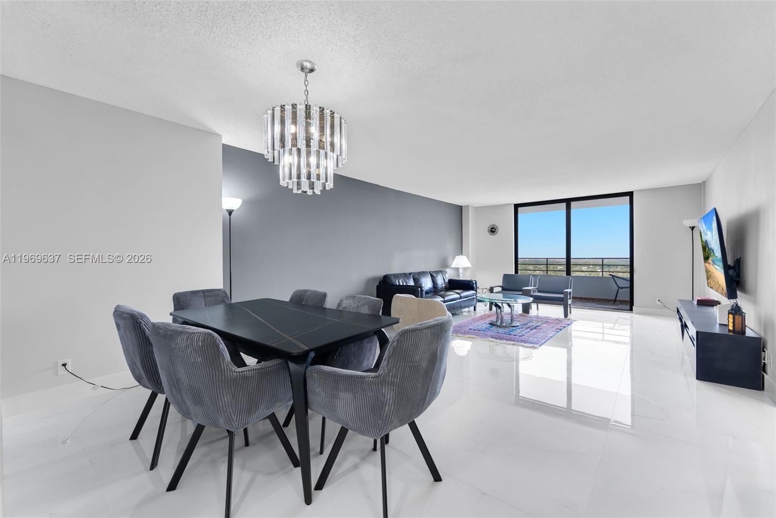 A modern living and dining area features white tile floors, a crystal chandelier, and balcony views — 2500 PARKVIEW DR # 2407, Hallandale Beach FL 33009, HALLANDALE BEACH, FL