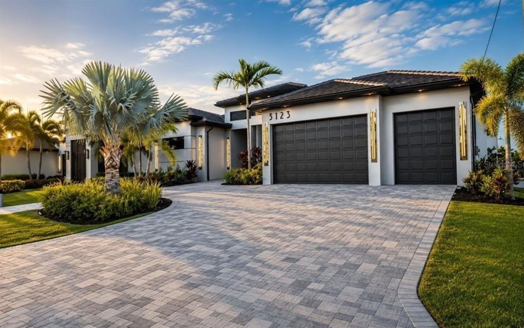 A modern house exterior features a paved driveway, dark garage doors, and surrounding palm trees — 3123 SW 29TH AVENUE, CAPE CORAL, FL