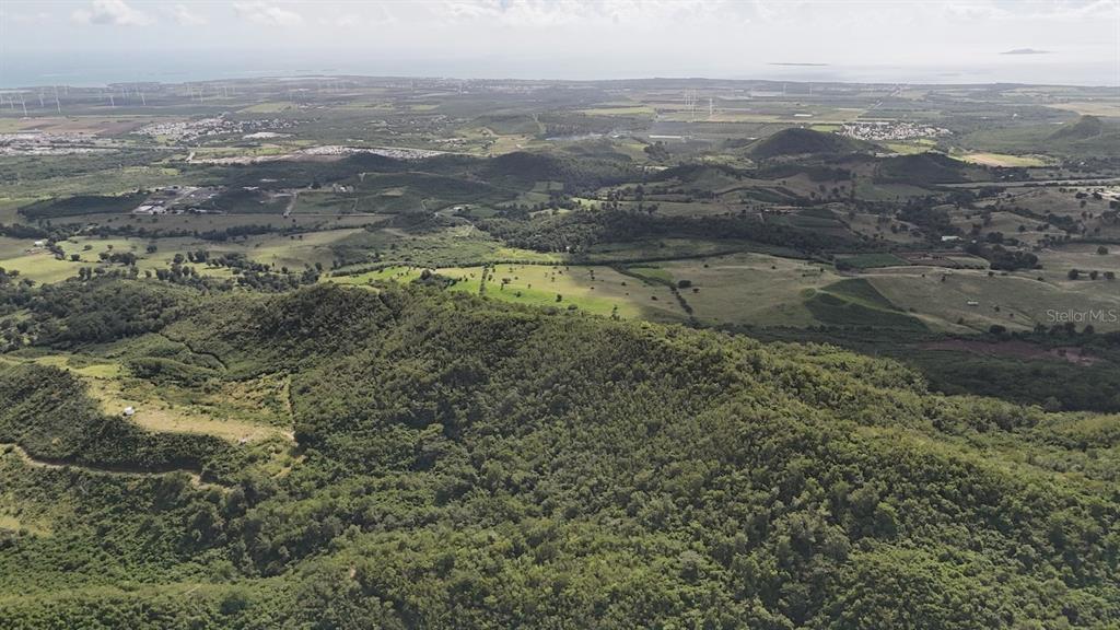 An aerial view shows a lush, hilly landscape with distant wind turbines and the ocean — 2565 PR545, COAMO, PR