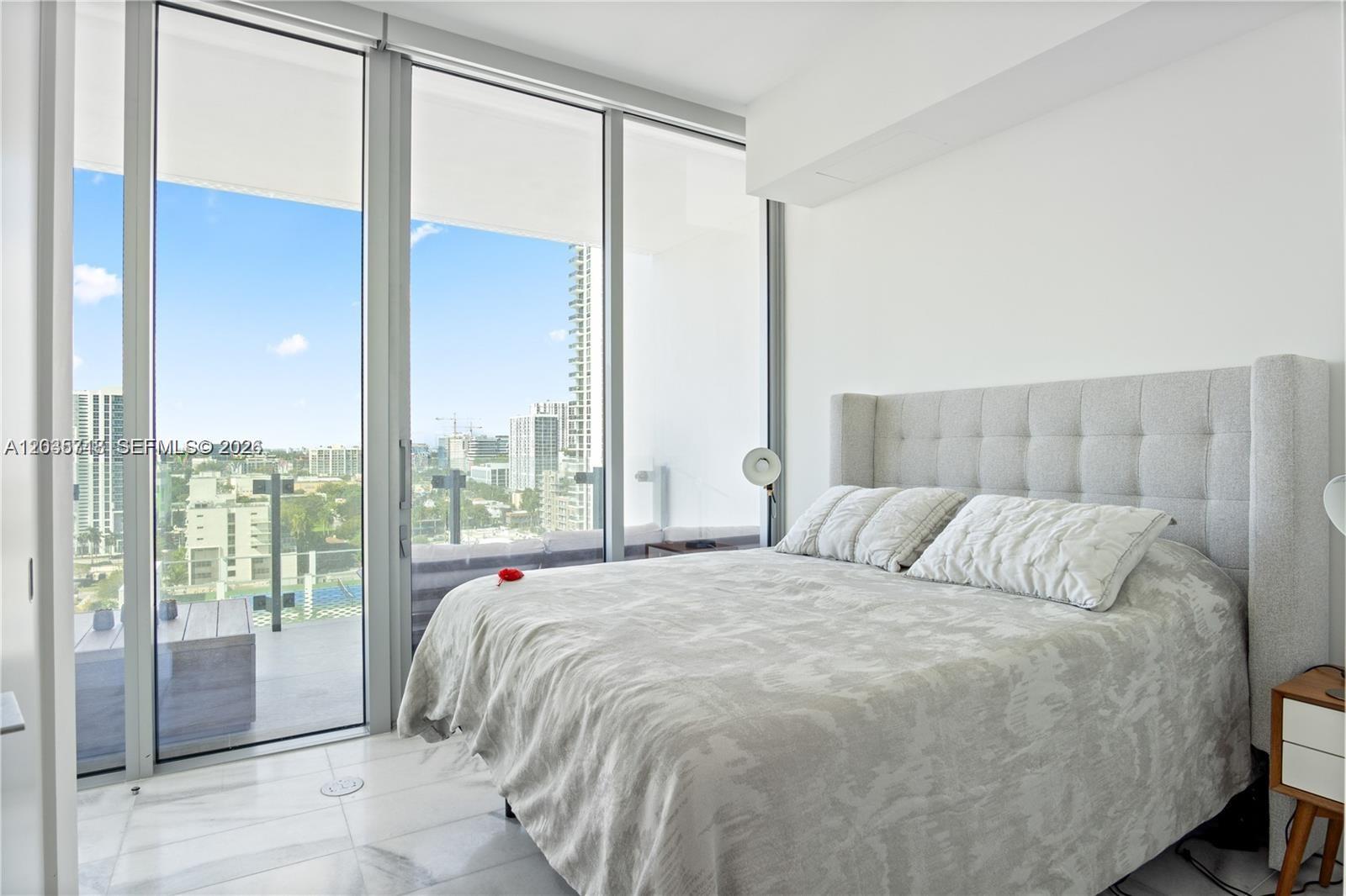 A bedroom features a tufted headboard, marble floors, and floor-to-ceiling windows overlooking a city skyline — 700 NE 26th Ter # 1405, Miami FL 33137, MIAMI, FL