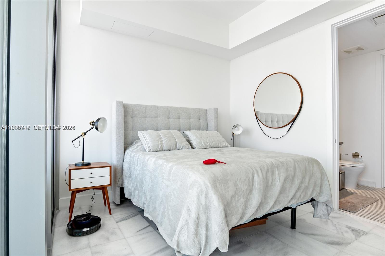 A bedroom features a gray tufted headboard, marble floors, a round mirror, and an attached bathroom — 700 NE 26th Ter # 1405, Miami FL 33137, MIAMI, FL