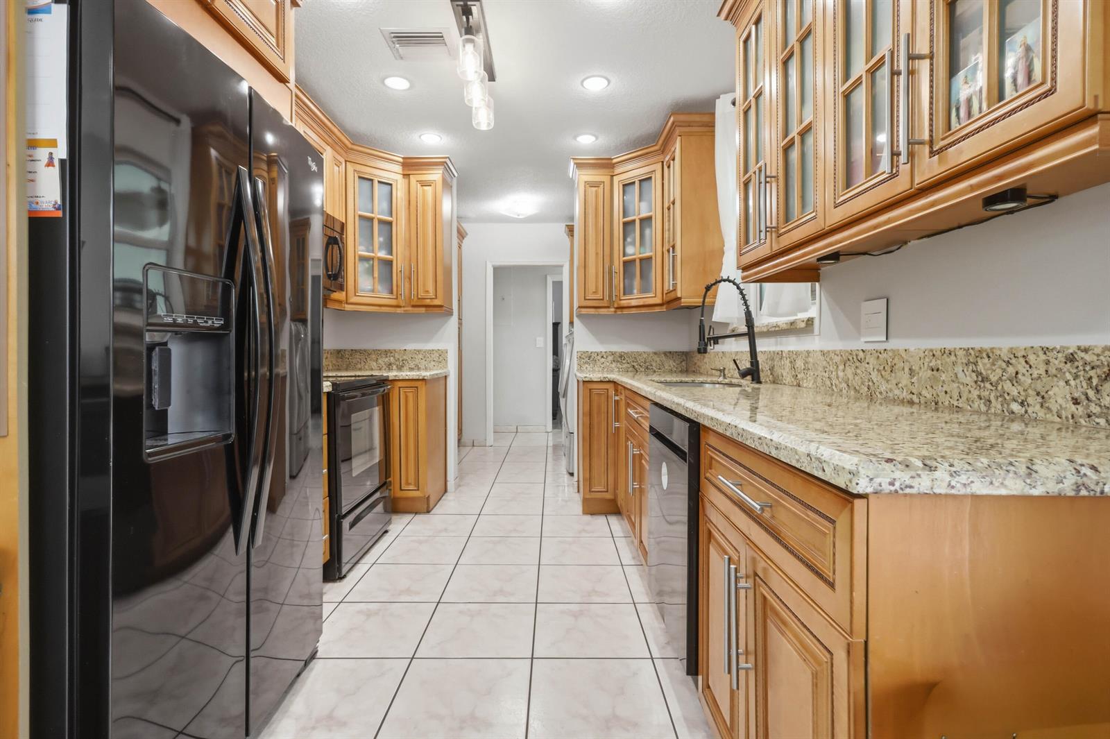 A galley kitchen features wood cabinets, granite countertops, black appliances, and light-colored tile flooring — 6448 Custer Street, Hollywood FL 33024, HOLLYWOOD, FL