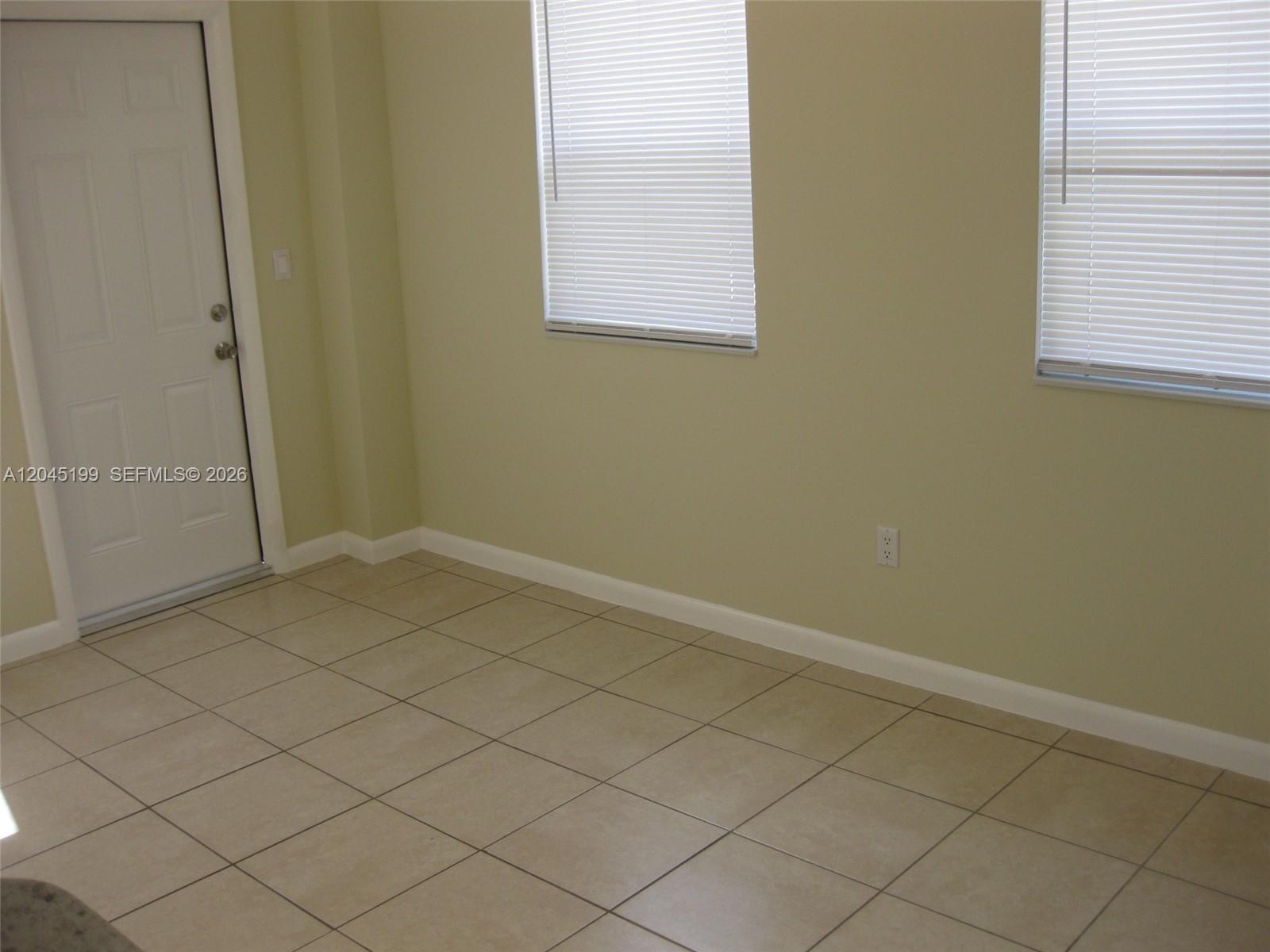 An empty room features beige walls, tiled flooring, two windows with blinds, and a door — 1520 NE 14th St, Homestead FL 33033, HOMESTEAD, FL