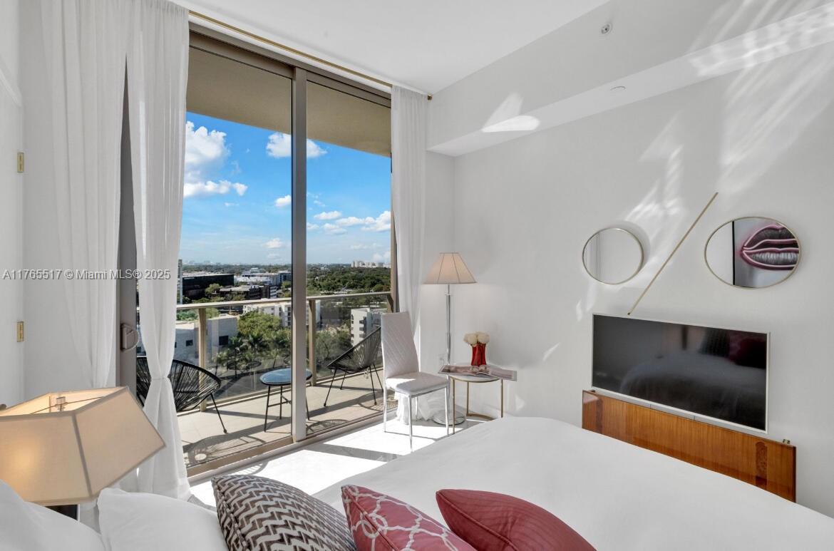 A bedroom features a white bed, a wall-mounted television, and a balcony with outdoor seating — 2678 Tigertail # 1009, Miami FL 33133, MIAMI, FL