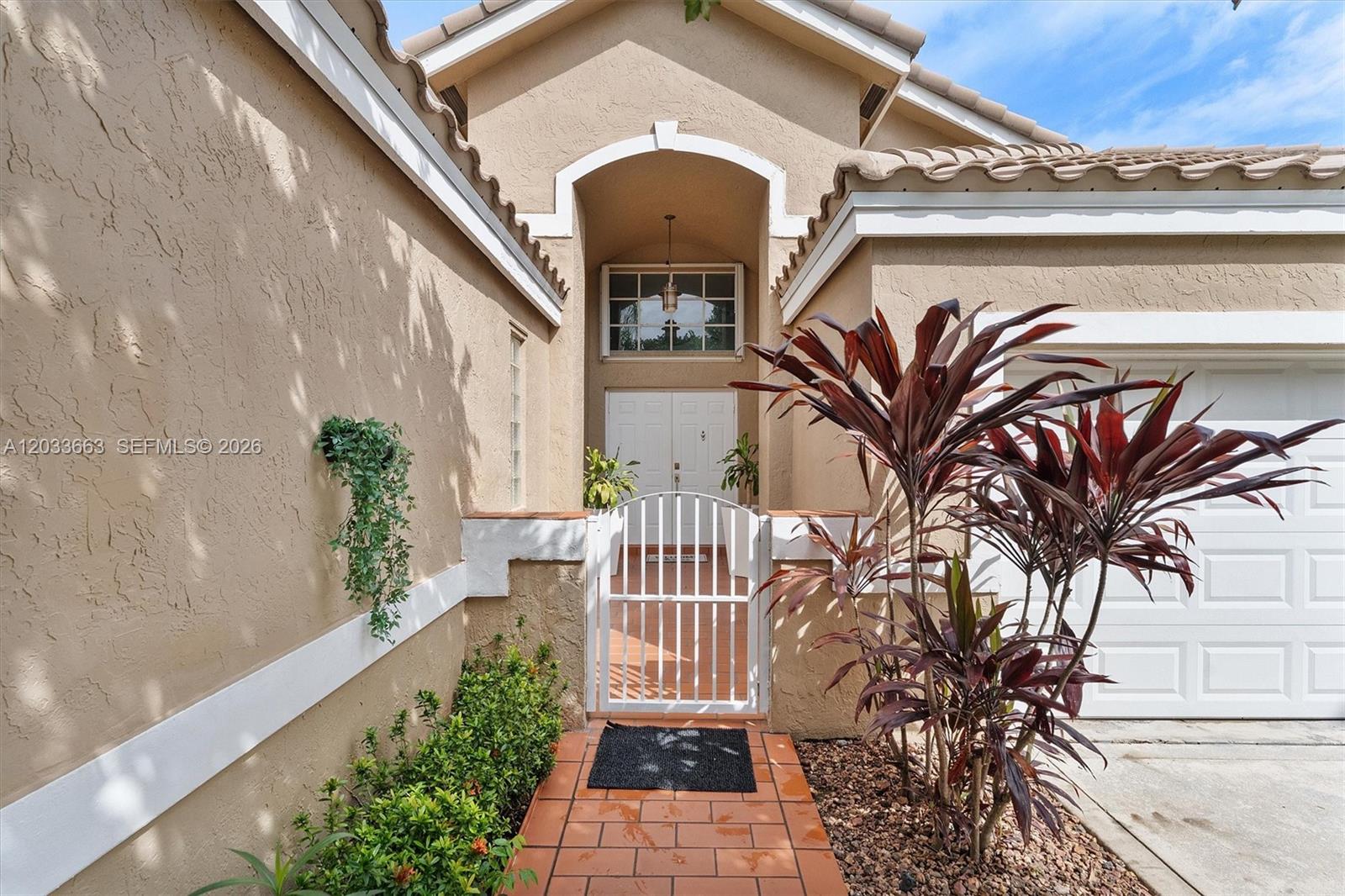 A tan stucco house features a gated courtyard entrance with a tiled walkway and landscaping — 2013 Island Cir, Weston FL 33326, WESTON, FL