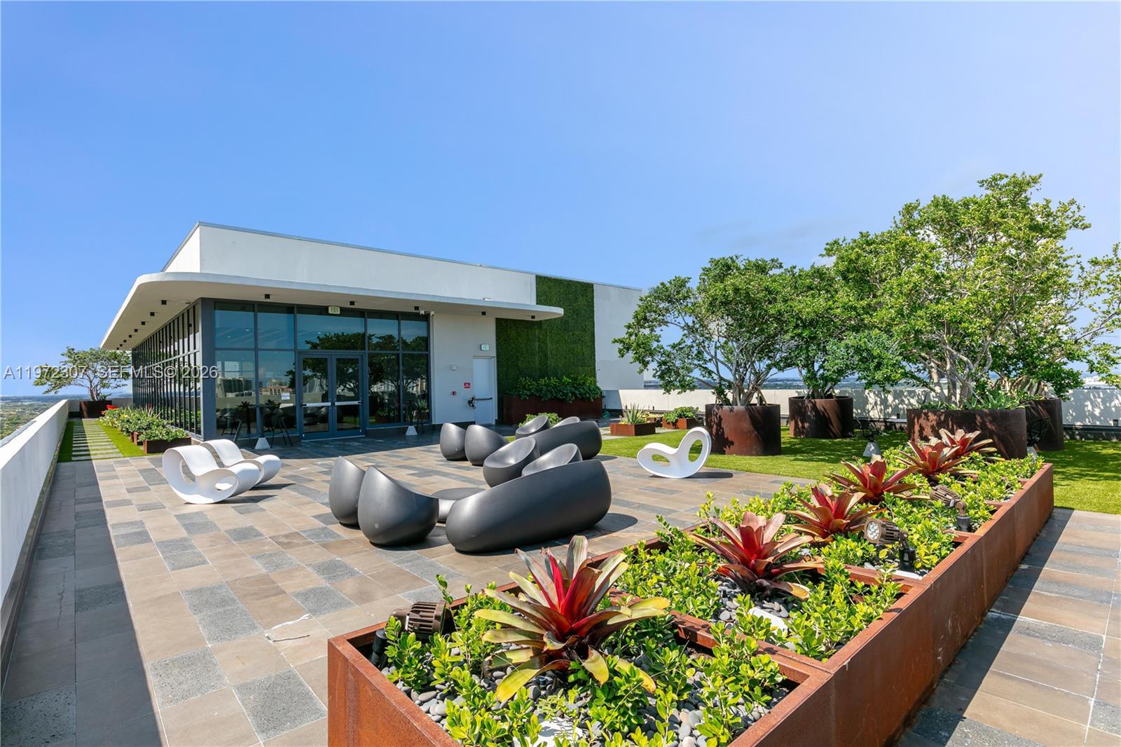 A rooftop terrace features modern outdoor furniture, planters with bromeliads, and trees under a clear sky — 19790 W Dixie Hwy 1004 Hwy, FL 33180, FL