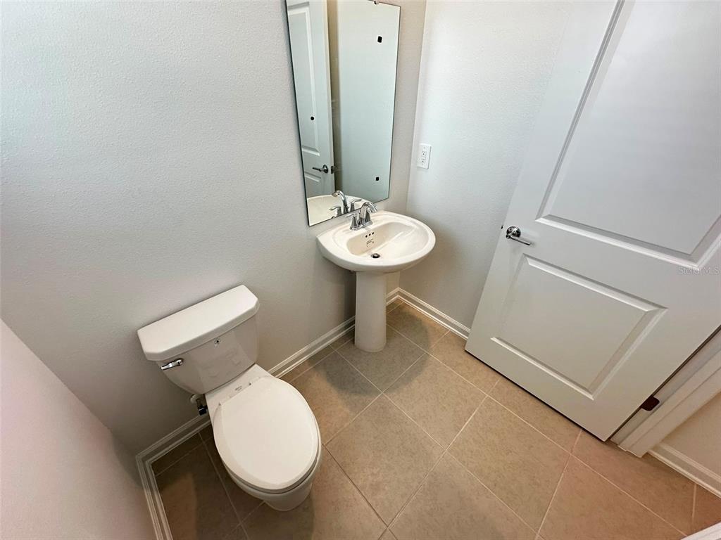 A small bathroom features a pedestal sink, a toilet, a mirror, and light-colored tile flooring — 6095 WELLBEING WAY, CLERMONT, FL