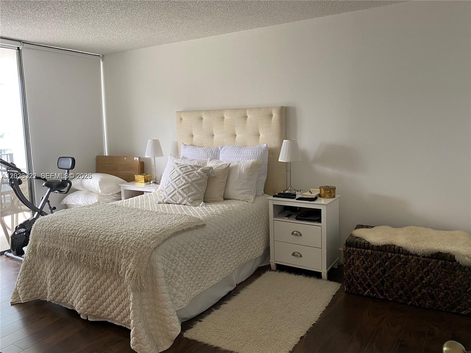A bedroom features a tufted headboard, white nightstands, a textured bedspread, and a stationary bike — MIAMI, FL