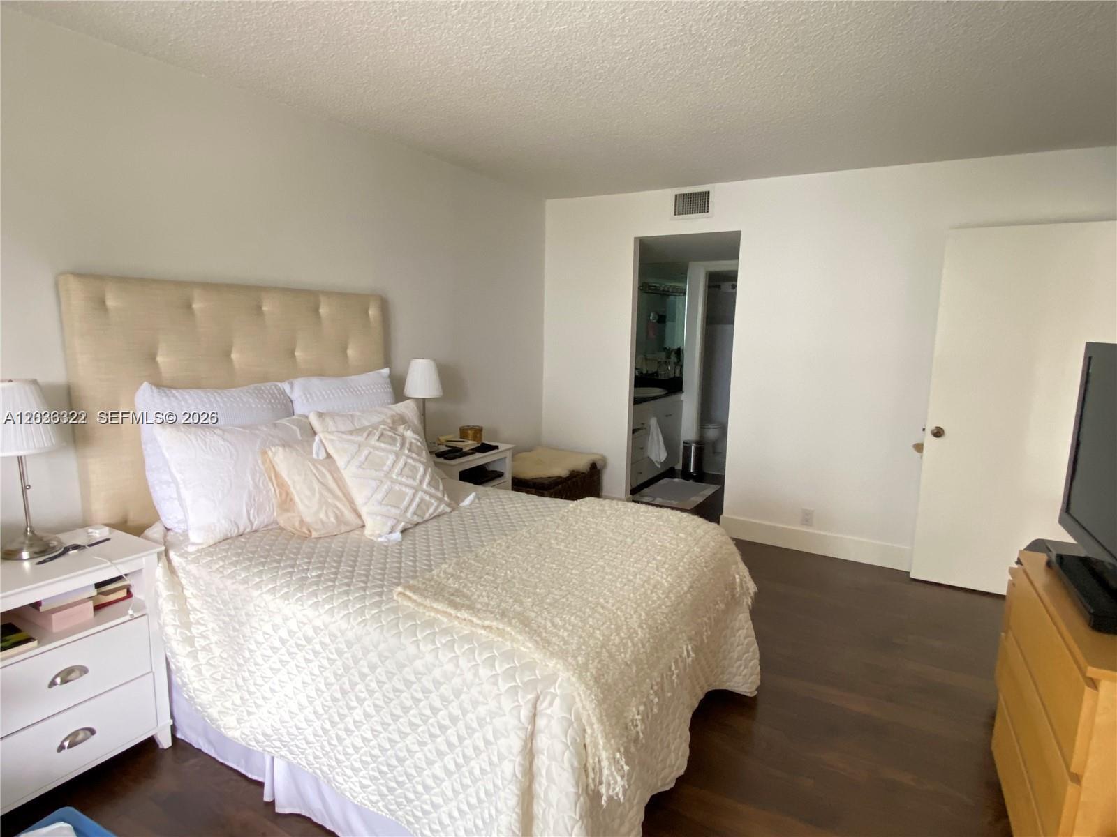 A bedroom features a tufted headboard, white nightstands, dark wood flooring, and an attached bathroom — MIAMI, FL