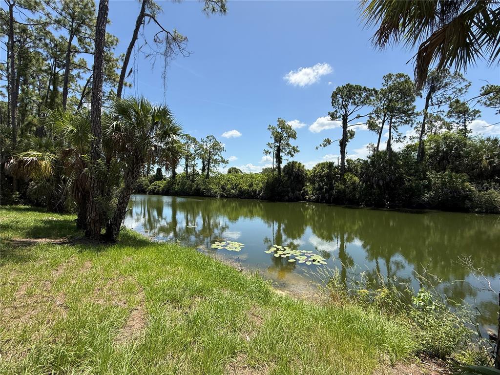 A grassy bank overlooks a calm pond surrounded by trees under a blue sky — 2646 SADNET LANE, NORTH PORT, FL