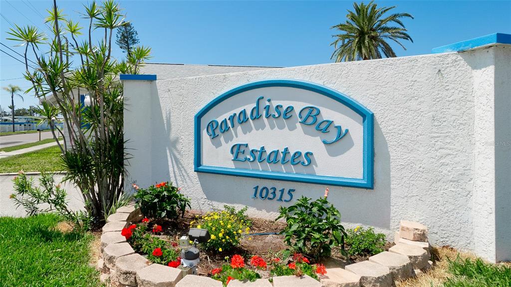A white stucco sign for Paradise Bay Estates stands in a landscaped garden bed — 10315 CORTEZ ROAD W UNIT 31-1, BRADENTON, FL