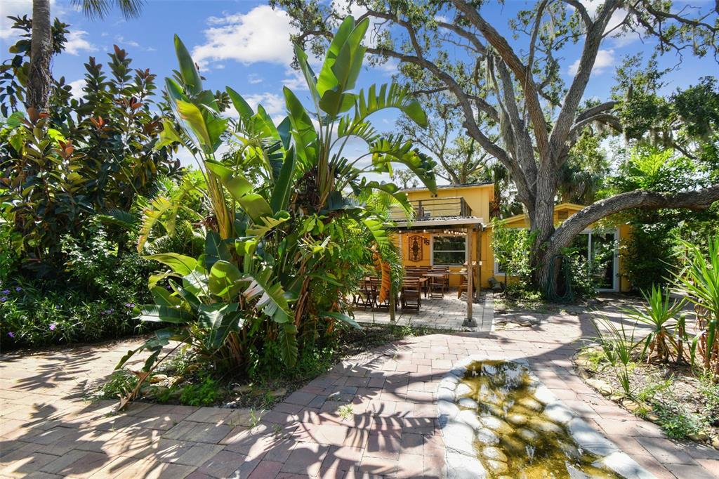 A brick patio features a small stone water feature, lush tropical plants, and a yellow house — 5909 MOHR ROAD, TAMPA, FL