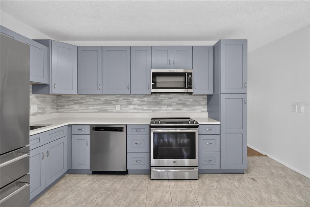 A kitchen features blue cabinets, stainless steel appliances, a tiled backsplash, and light-colored floor tiles — 2295 AMERICUS BOULEVARD E UNIT 59, CLEARWATER, FL