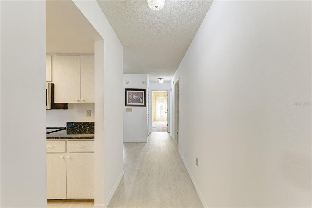 A hallway with light wood-look flooring leads toward a kitchen and a distant sunlit room — 804 67TH STREET W, BRADENTON, FL