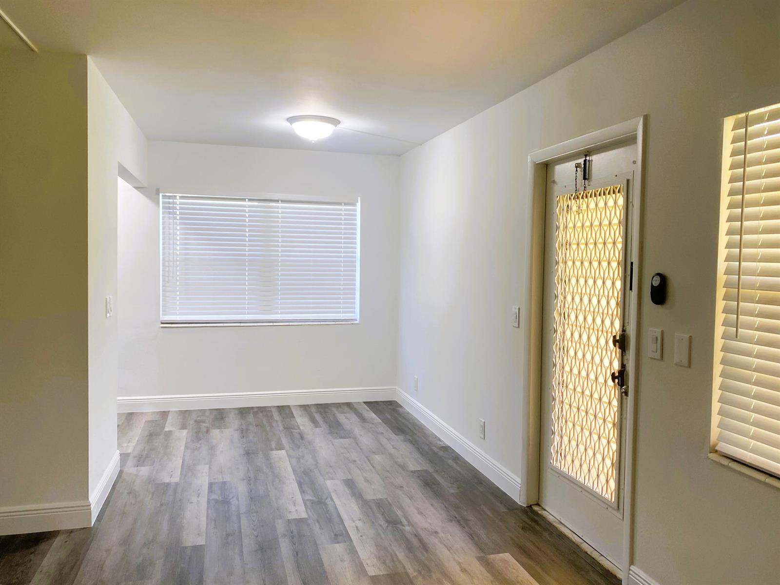 An empty room features white walls, gray wood-look flooring, two windows with blinds, and a door — 256 Mansfield # G, Boca Raton FL 33434, BOCA RATON, FL