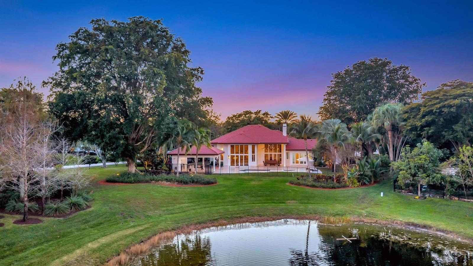A house with a red tile roof sits behind a pond and large trees — 3460 Windmill Ranch Road, Weston FL 33331, WESTON, FL