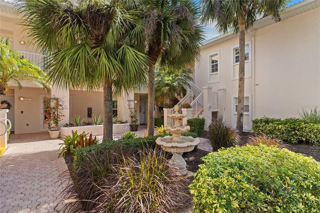 A courtyard with a stone fountain, palm trees, and a two-story building with beige stucco — 4244 CENTRAL SARASOTA PARKWAY UNIT 716, SARASOTA, FL