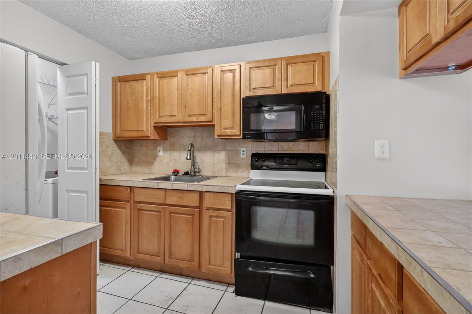 A kitchen features light wood cabinets, black appliances, tile countertops, and white floor tiles — 20731 NE 4th Pl # 104, Miami FL 33179, MIAMI, FL