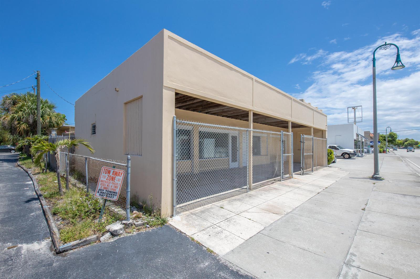 A tan commercial building with a chain-link fence sits along a sidewalk under a blue sky — 922 S Dixie Highway, FL 33460, FL