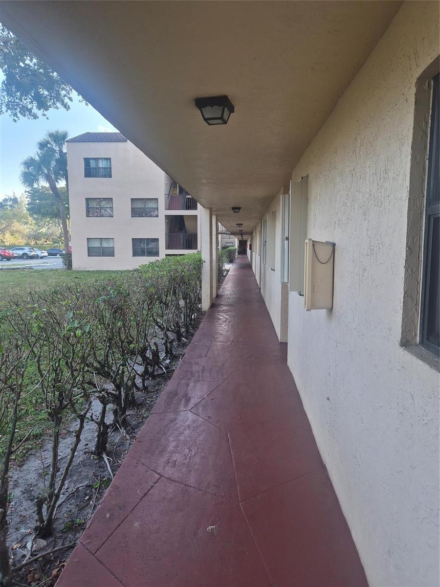 An outdoor covered walkway with red painted flooring runs alongside a hedge and building exterior — 3252 Foxcroft Road # 113, Miramar FL 33025, MIRAMAR, FL
