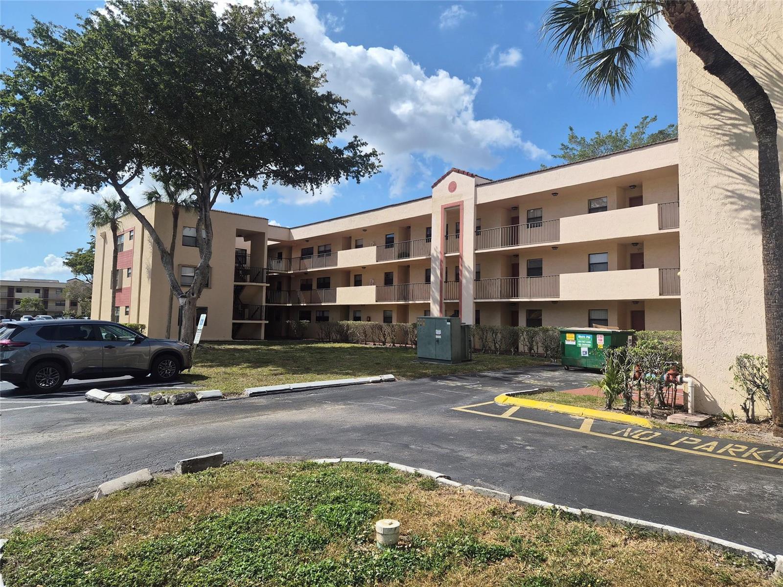 An exterior view of a multi-story apartment building with a parking lot and dumpster — 3252 Foxcroft Road # 113, Miramar FL 33025, MIRAMAR, FL