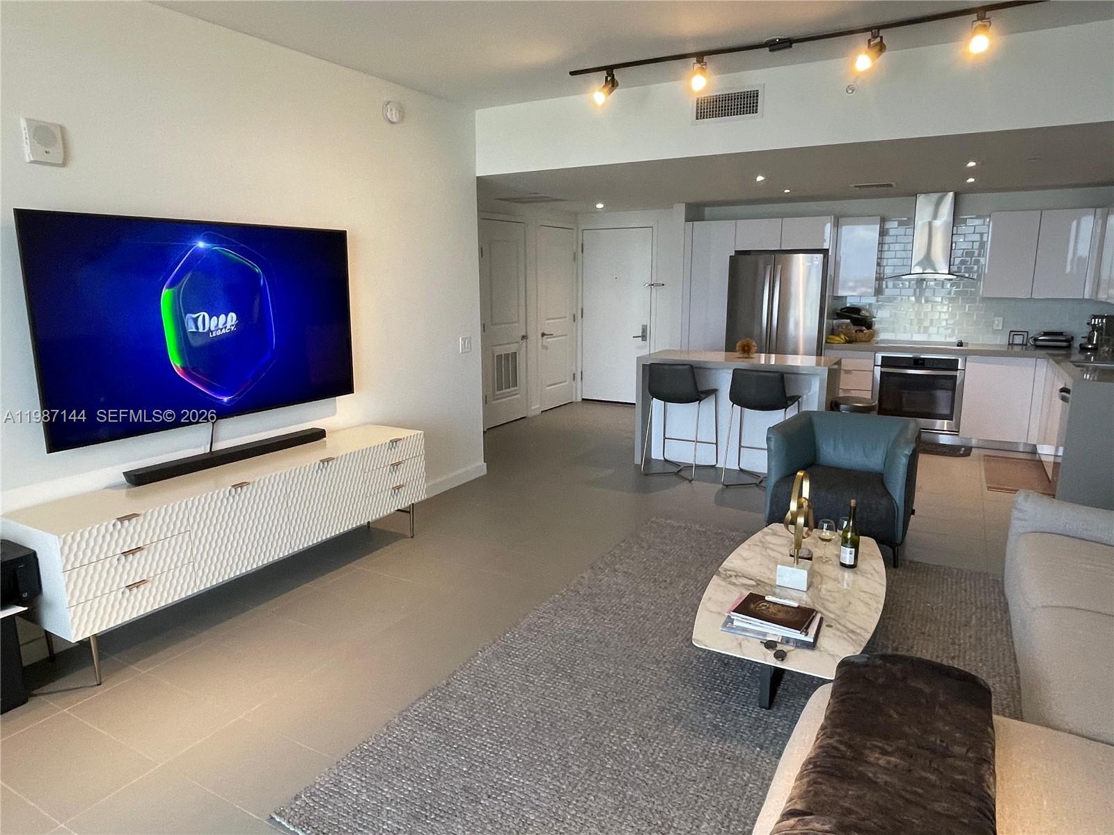 A modern living room features a large television, white media console, and an open kitchen — 1600 NE 1st Ave # 3717, Miami FL 33132, MIAMI, FL