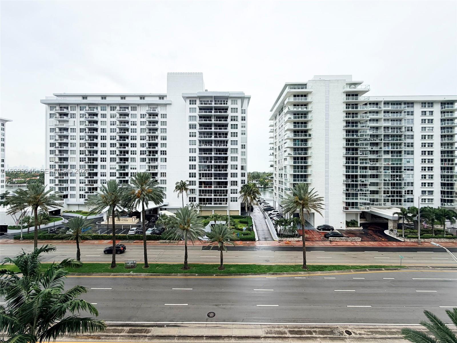 An exterior view shows two tall white apartment buildings separated by a driveway and palm trees — 5757 Collins Ave # 702, Miami Beach FL 33140, MIAMI BEACH, FL