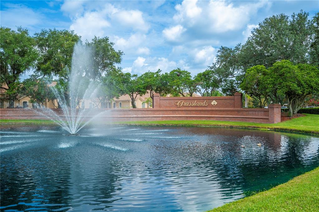 A brick wall with the Grasslands sign stands behind a pond with a water fountain — 684 GRASSLANDS VILLAGE CIRCLE, LAKELAND, FL