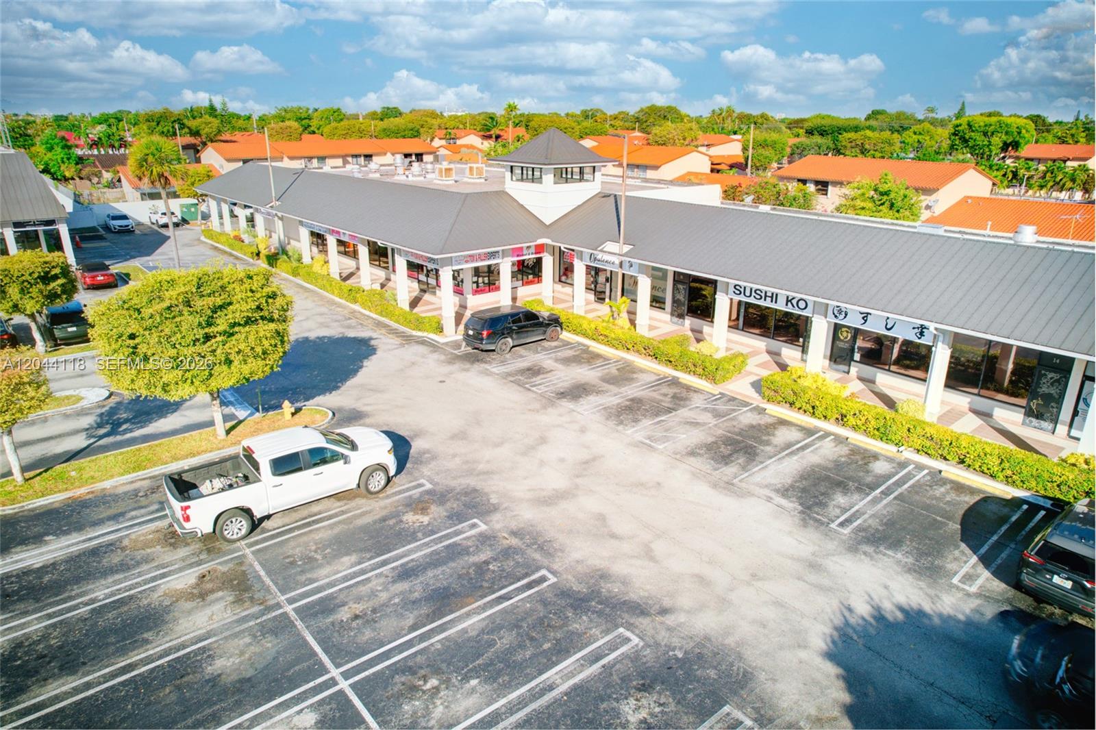 An aerial view shows a commercial strip mall with a parking lot and surrounding residential homes — MIAMI, FL
