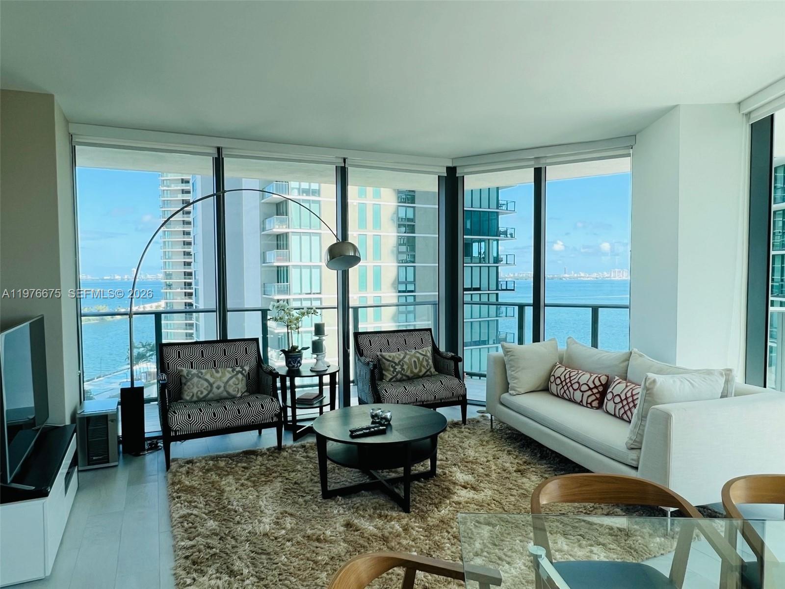 A living room with floor-to-ceiling windows, a white sofa, patterned chairs, and water views — 501 NE 31st St # 1606, Miami FL 33137, MIAMI, FL
