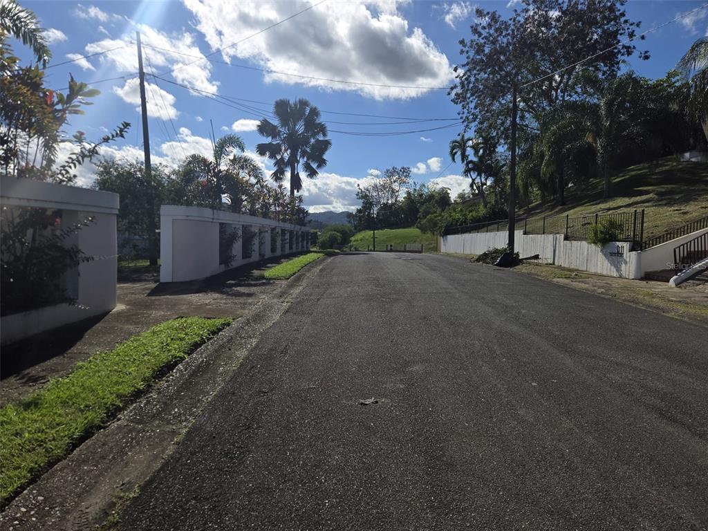A paved road leads uphill between white concrete walls and lush tropical landscaping — E-4 URB. EL RETIRO CALLE RECREO, CAGUAS, PR