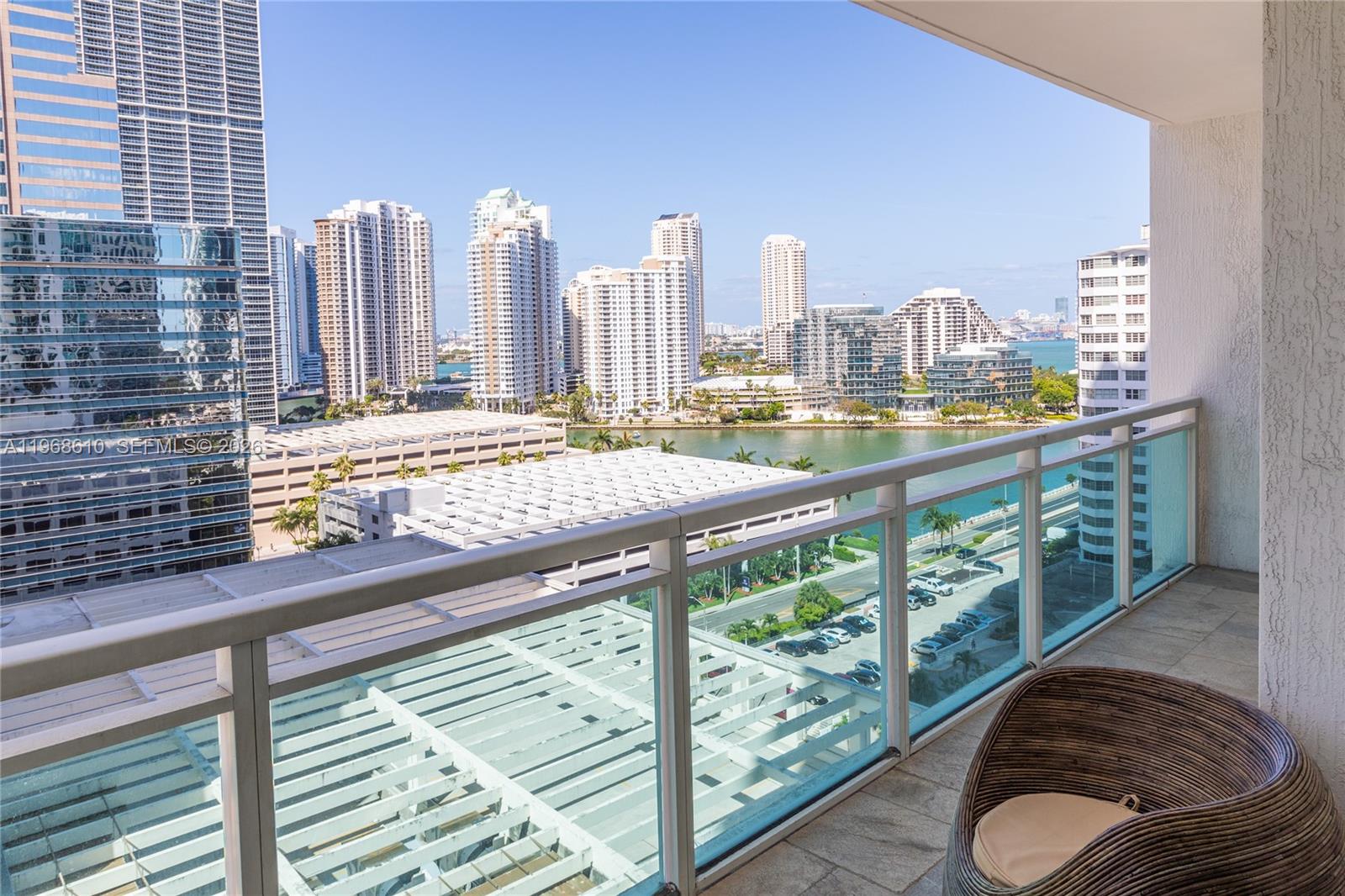 A balcony with a wicker chair overlooks a city skyline, a waterway, and parking structures — 950 Brickell Bay Dr # 1504, Miami FL 33131, MIAMI, FL
