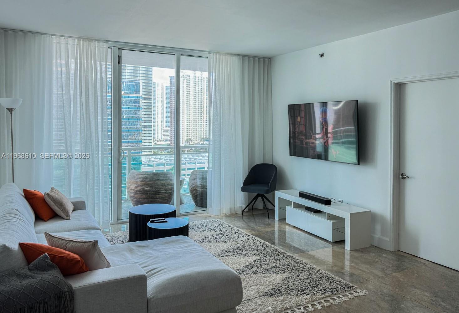 A living room features a white sofa, a television, and a balcony overlooking city buildings — 950 Brickell Bay Dr # 1504, Miami FL 33131, MIAMI, FL